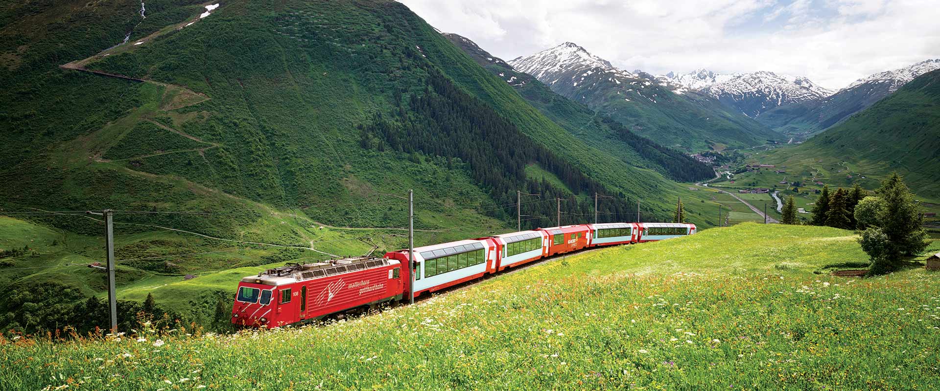 Red train in green pastures with snow capped mountains in background